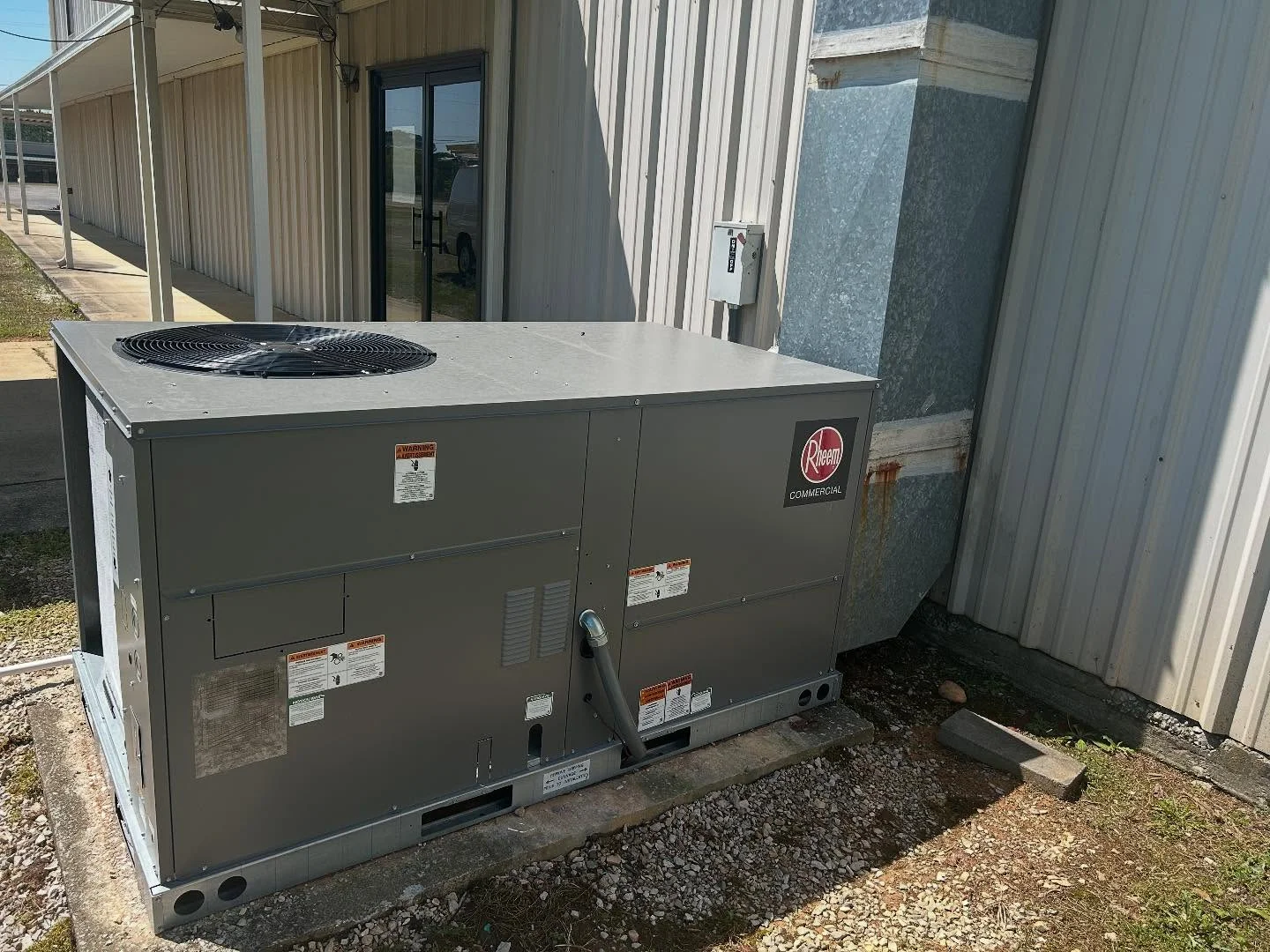 Rheem commercial heat pump unit installed on gravel pad beside metal building with dual fans and electrical connections